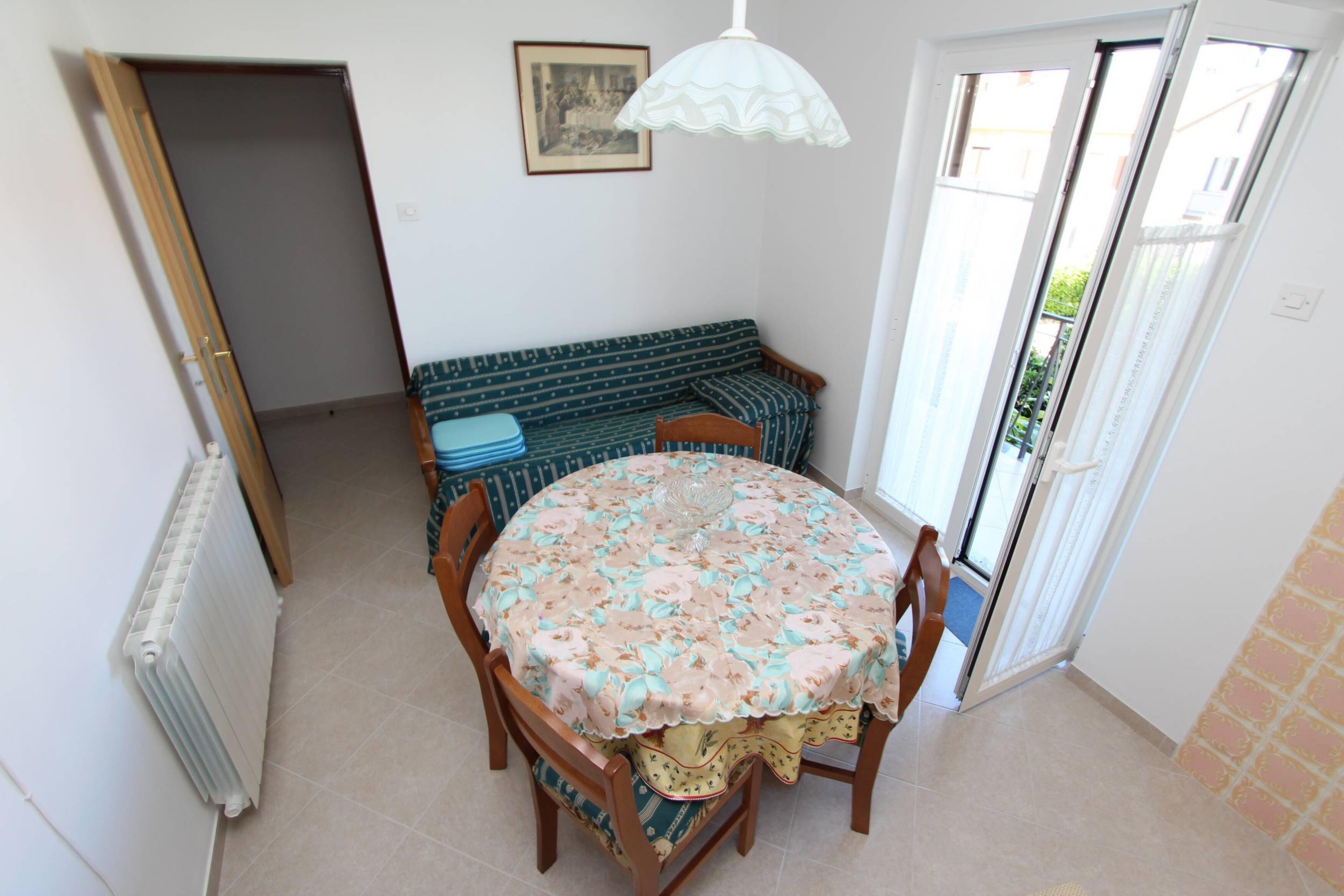 A dining area with a round table, chairs, a patterned tablecloth, a bench, and a door leading outside.