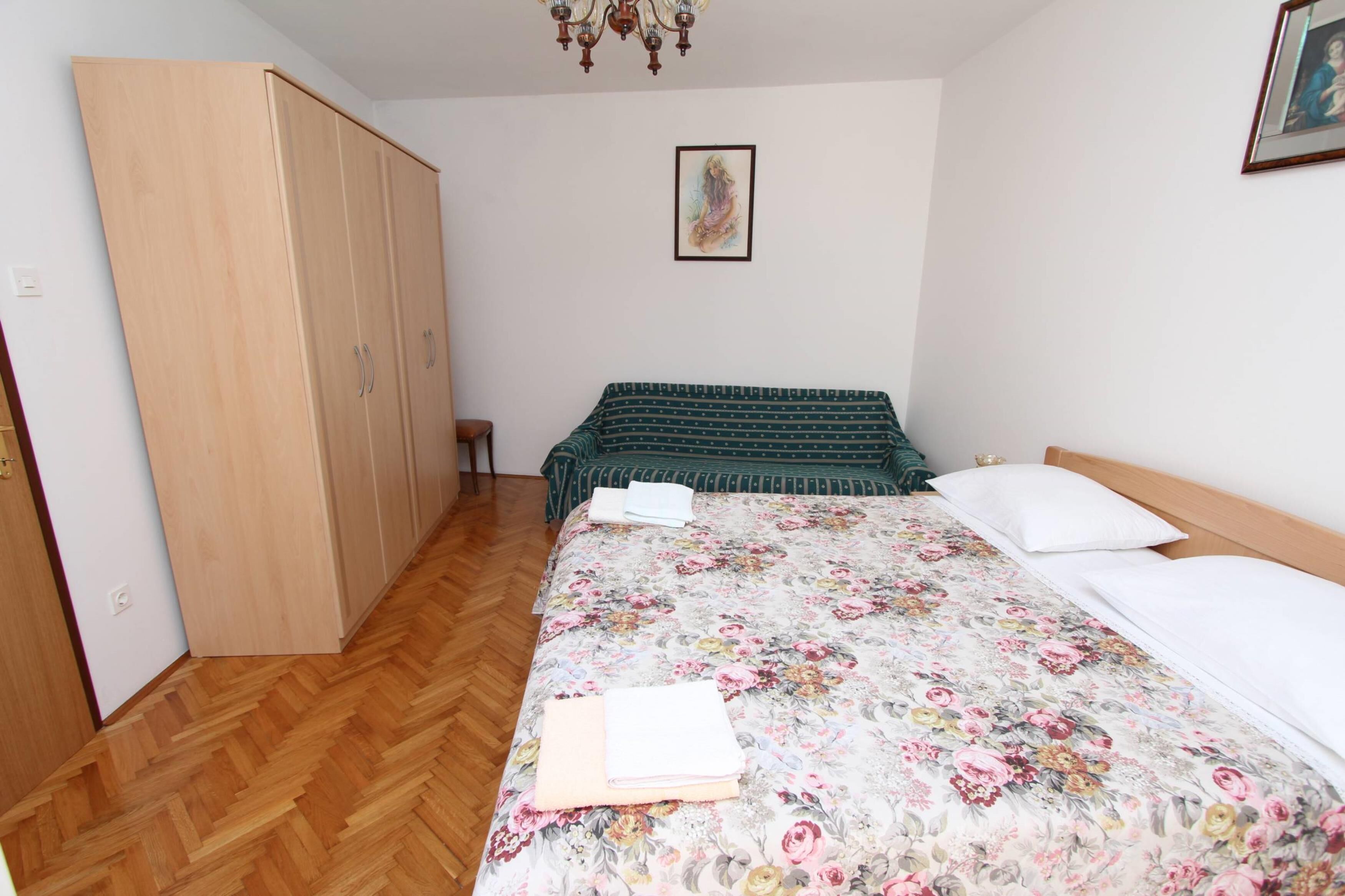A bedroom with a wooden wardrobe, a bed with floral bedding, and a small table with a book.