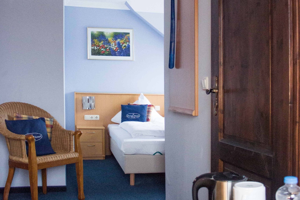 A hotel room with a bed, a chair, a nightstand, a painting on the wall, and a door.