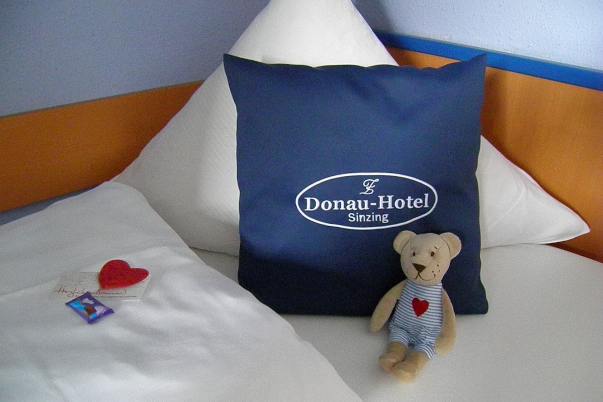 A bed with a pillow labeled 'Donau-Hotel Sinsing,' a teddy bear, and a heart-shaped object.