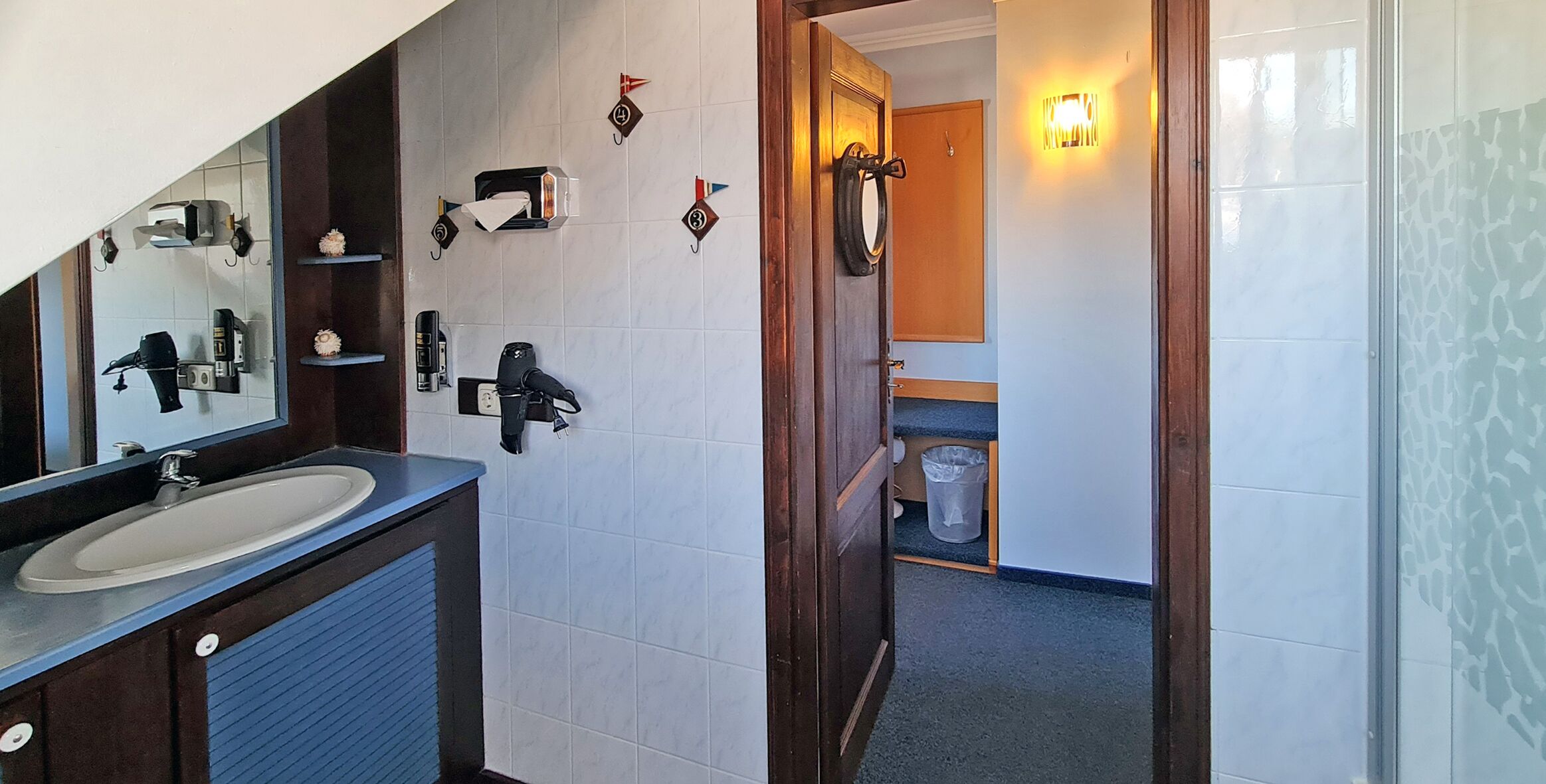 Un baño con lavamanos, espejo y perchero. Hay una puerta que da a otra habitación.