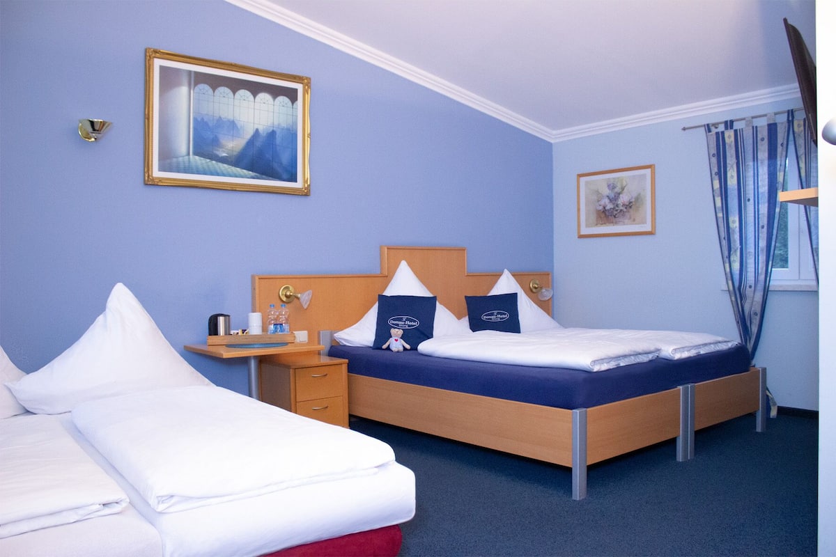 A hotel room with a bed, a desk, and a nightstand. There is a painting on the wall and a window with curtains.