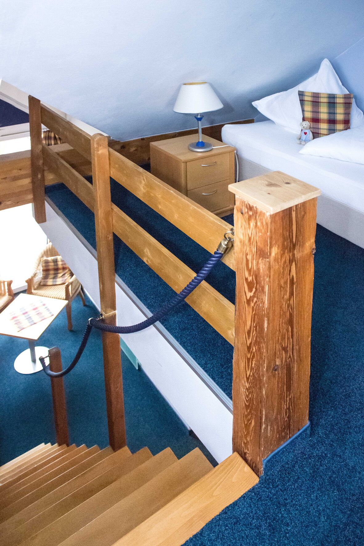 A bedroom with a wooden bunk bed, a bedside table with a lamp, a small bedside table, a bed with a plaid pillow, and a chair with a plaid cushion.