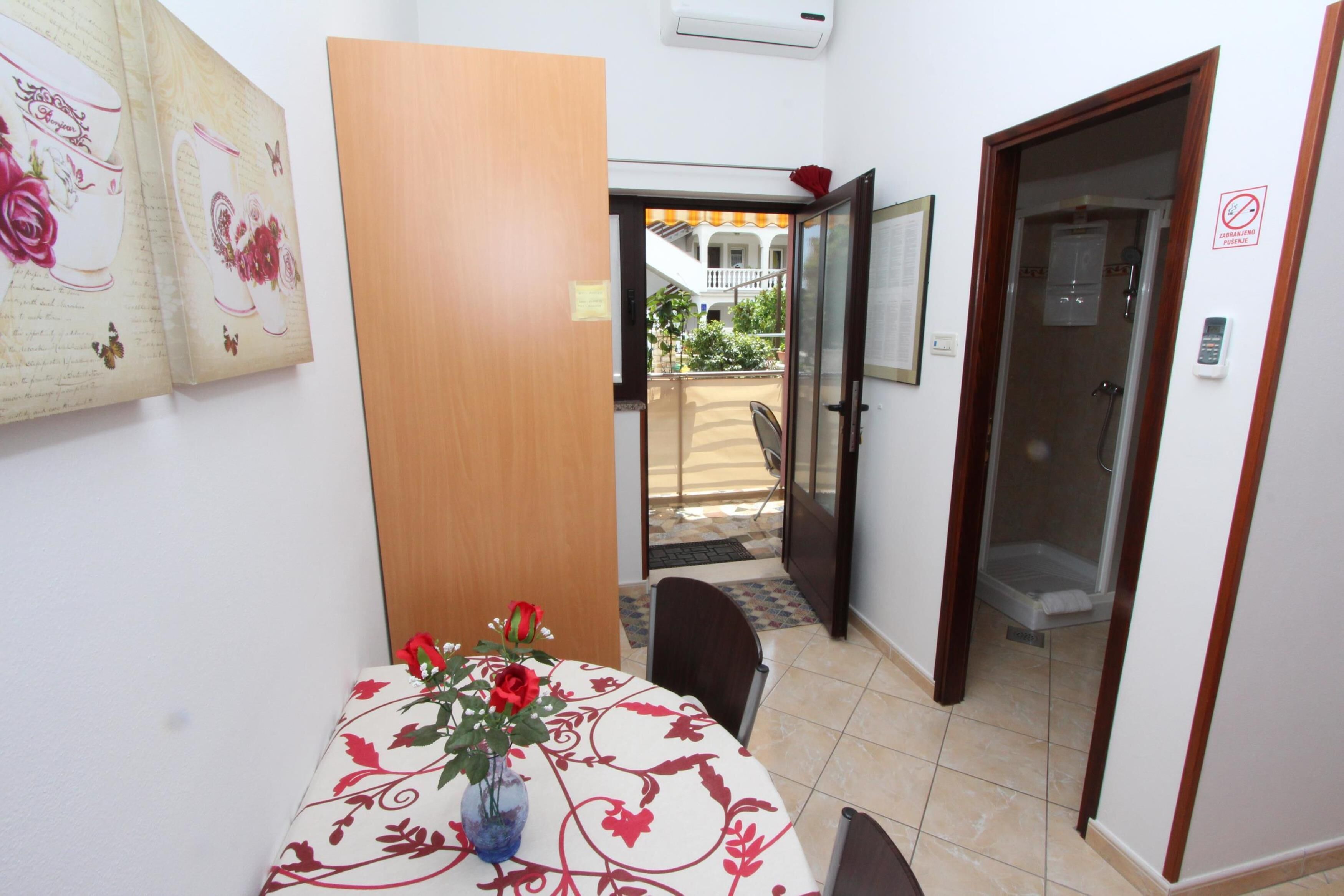A room with a dining table, a vase with flowers, a wooden door, and a view of an outdoor balcony.