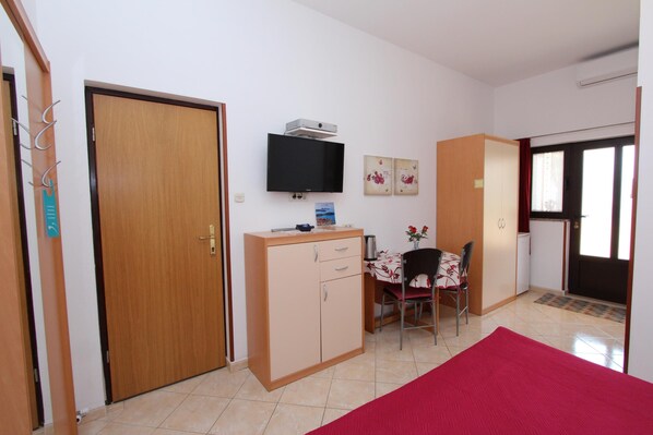 Una habitación pequeña y limpia, con una puerta de madera, un televisor fijado a la pared, una mesita con una silla y una alfombra roja en el suelo.