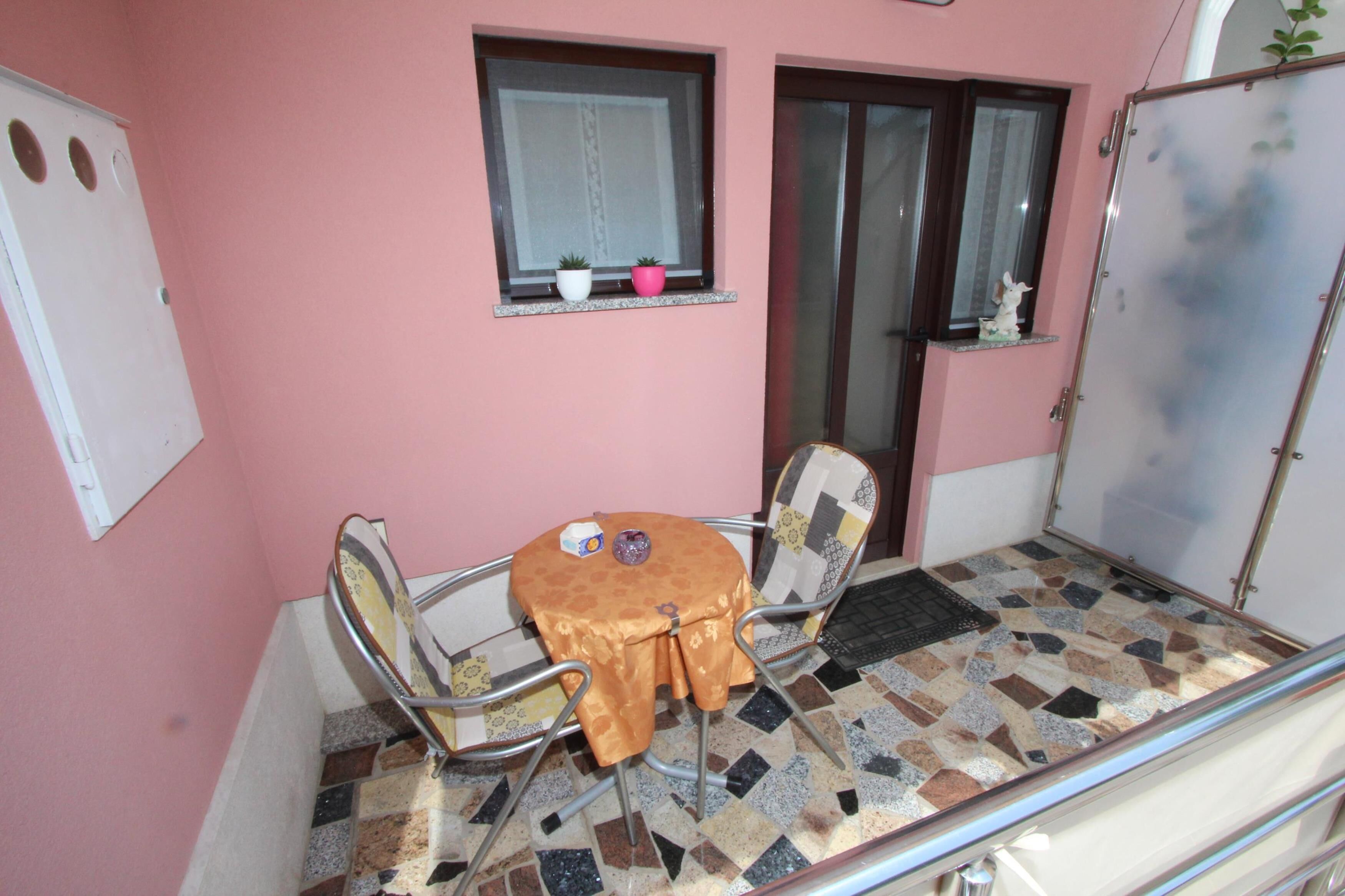 A small room with a pink wall, a tiled floor, a round table with two chairs, a window with a curtain, and a door with a mirror.