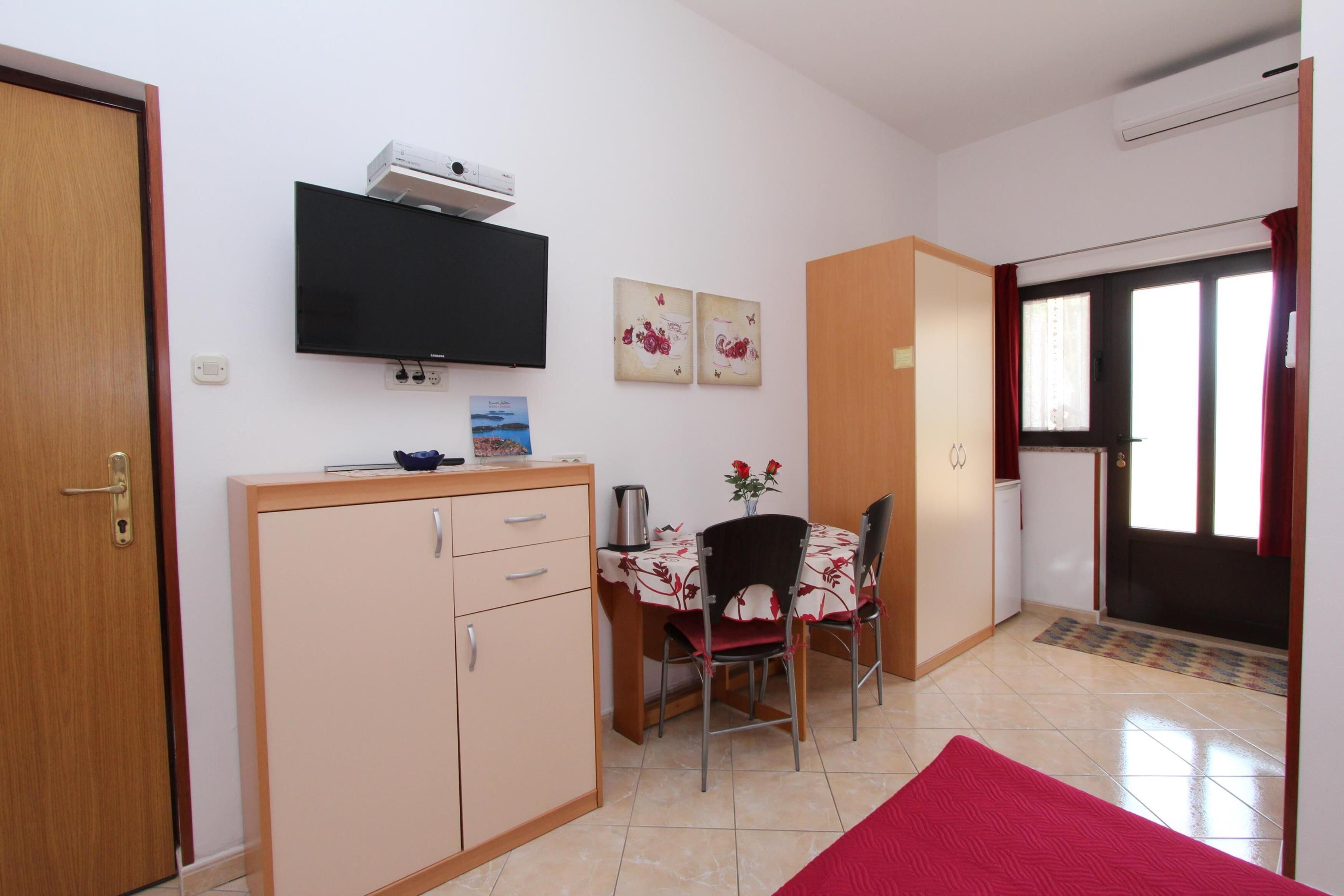 Een compacte kamer met een kitchenette, een eethoek met tafel en stoelen, een wandgemonteerde flatscreen-tv en een deur naar een andere kamer.