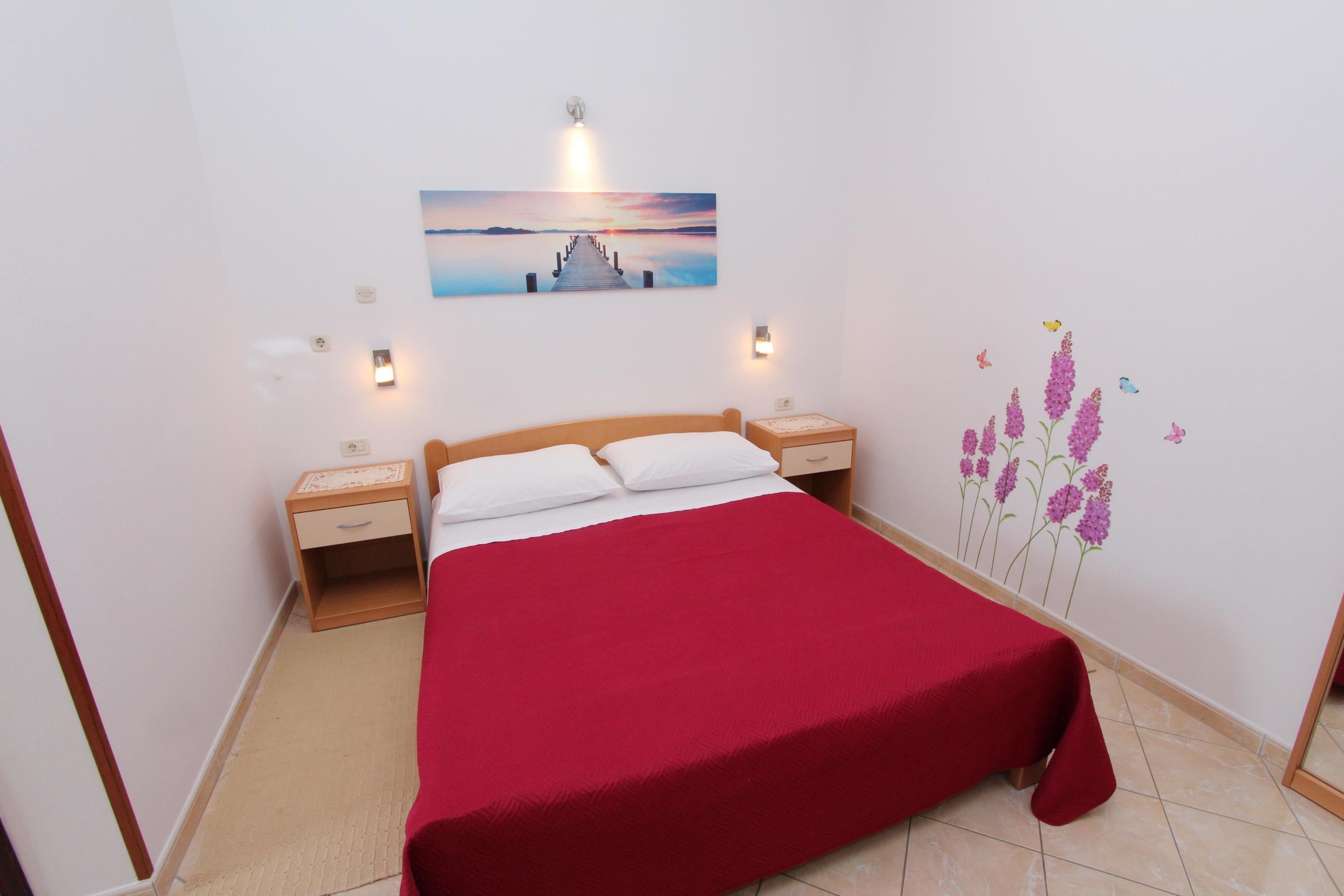 Ein Schlafzimmer mit einem hölzernen Bett, einer roten Bettdecke, zwei Nachttischen, einem Wandbild und einer Blumen-Wandsticker.
