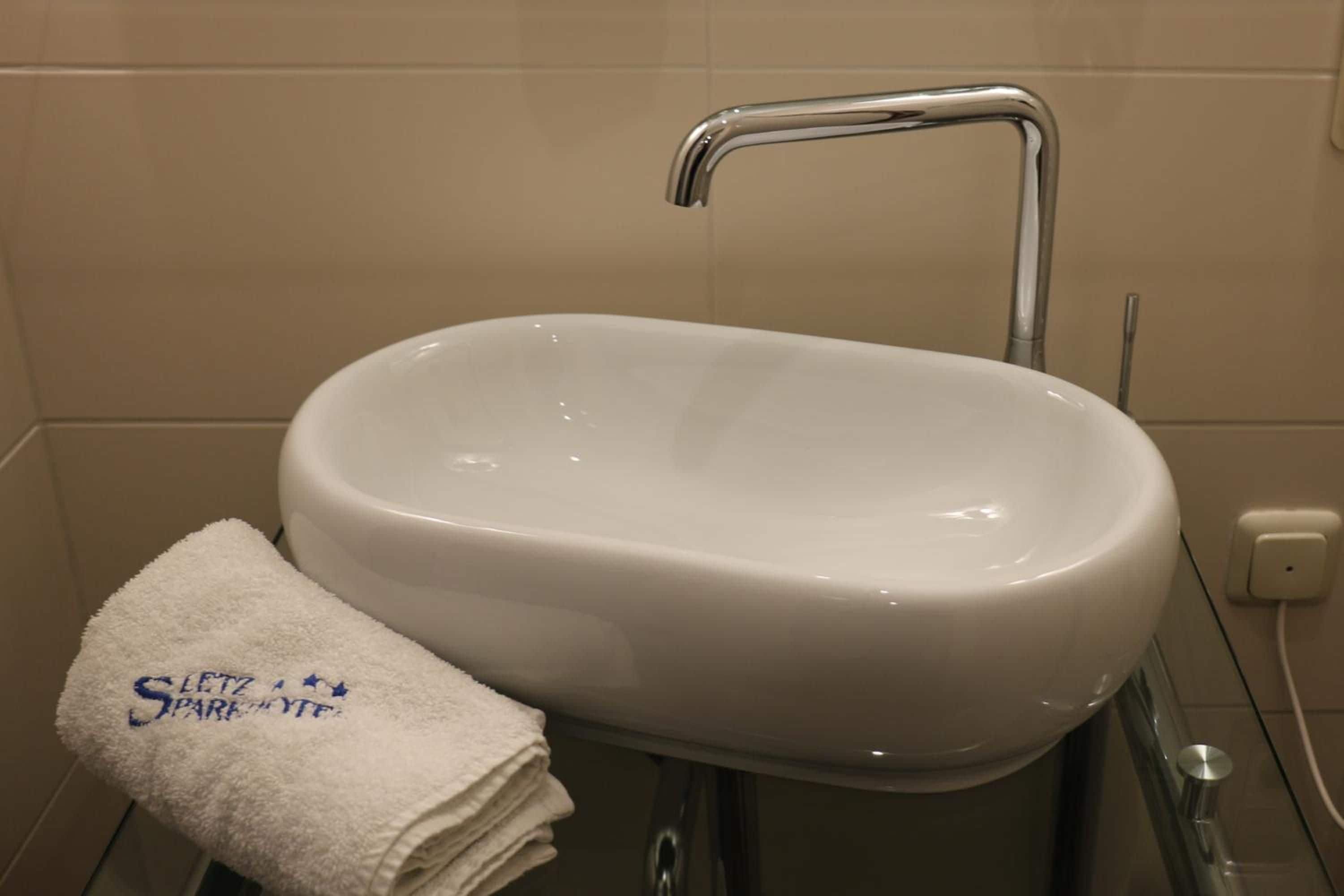 Ein weißes Waschbecken mit chromfarbenem Wasserhahn, ein weißes Handtuch mit Aufschrift und eine Steckdose an der Wand.