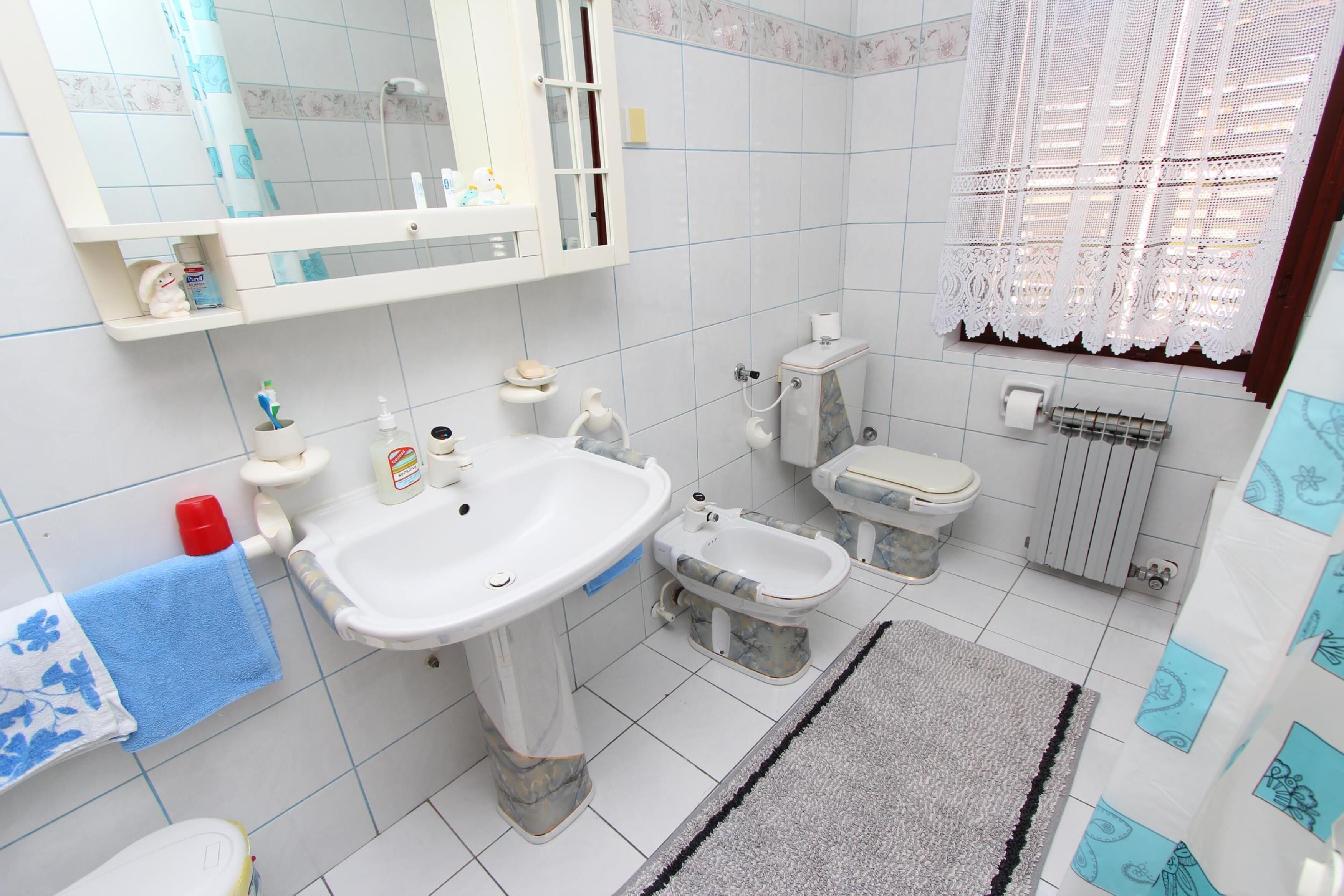 A bathroom with a sink, toilet, bidet, and a window with lace curtains.
