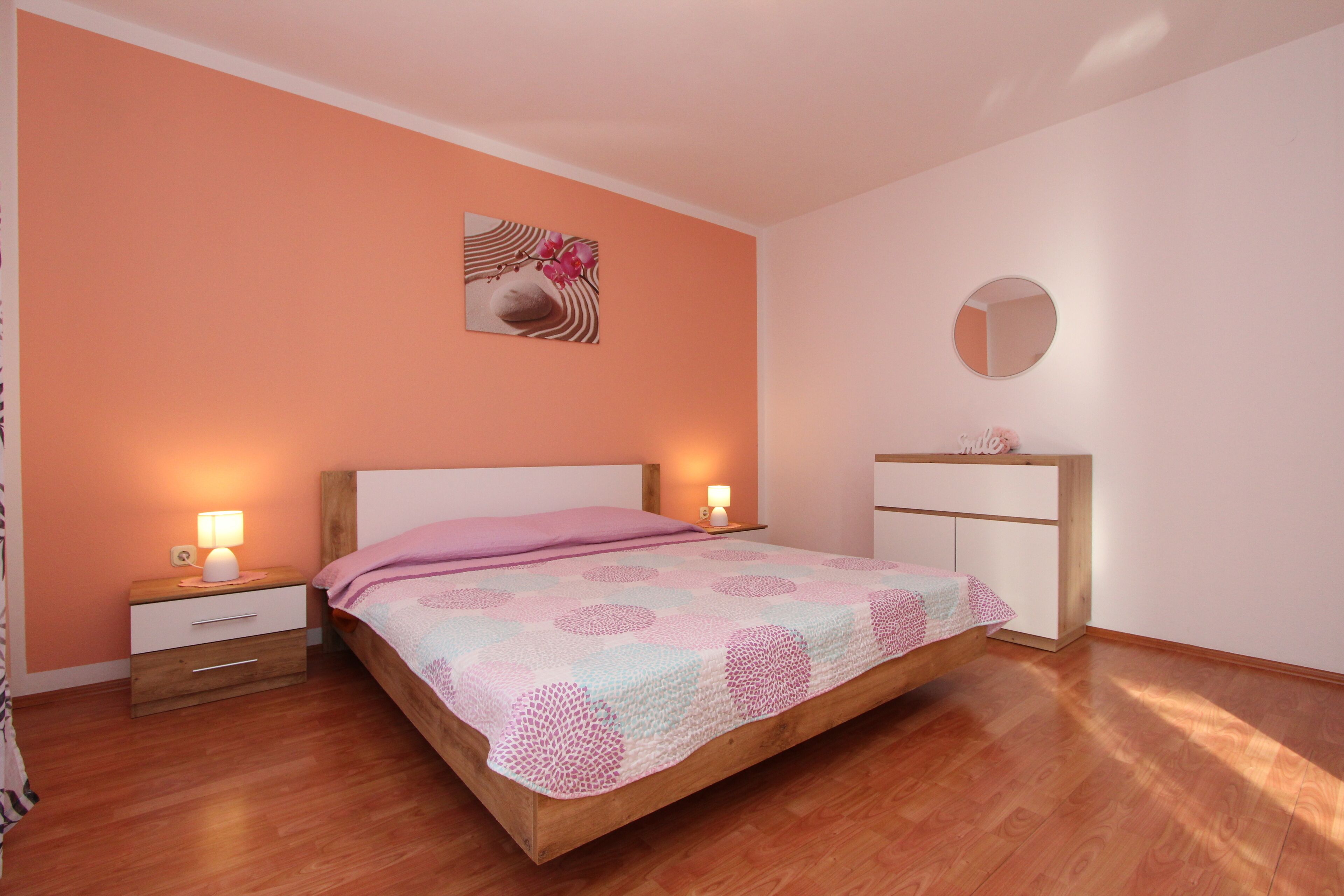 A bedroom with a wooden bed, two bedside tables with lamps, a dresser, and a round mirror on the wall.