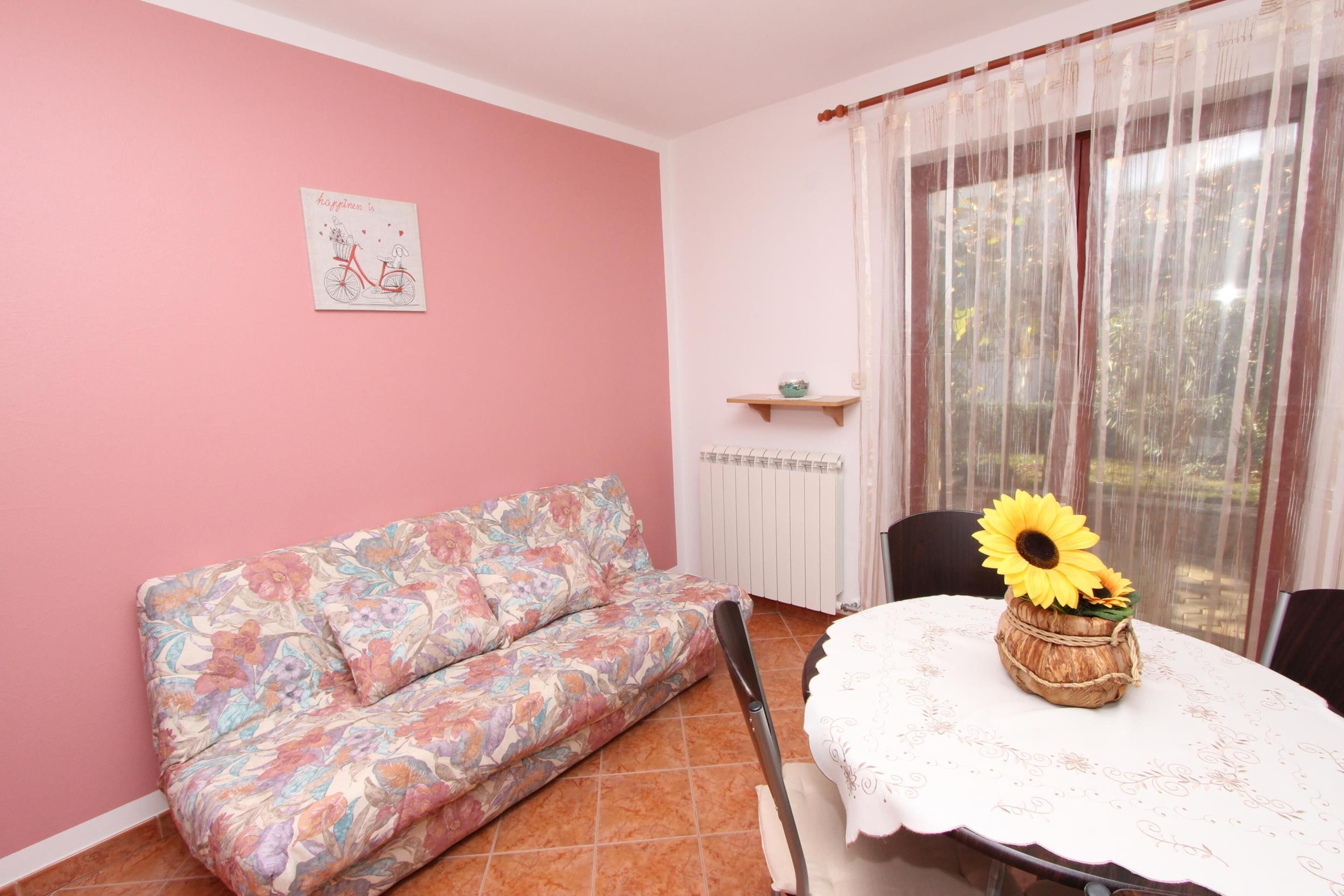 A room with a pink wall, a floral sofa, a table with a sunflower vase, and a window with sheer curtains.