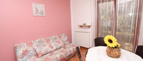 A room with a pink wall, a floral sofa, a table with a sunflower vase, and a window with sheer curtains.