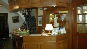 A wooden reception desk with a lamp, framed pictures, and a staircase in the background. - Kluster Hof (Basdahl)