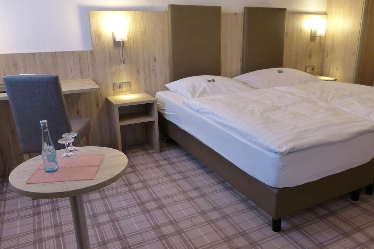 Hotellihuone, jossa on parivuode, pieni yöpöytä, tuoli ja pyöreä pöytä, jonka päällä on pullo ja laseja.