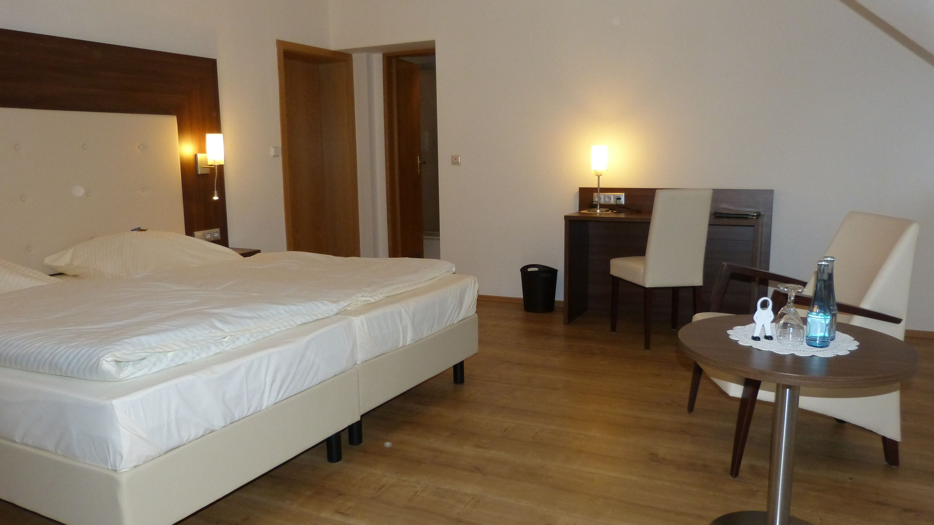 A hotel room with a bed, a desk, a chair, and a small table with a bottle and a glass.