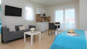 Una habitación de hotel moderna con sofá, una mesita con un florero, una cama con colcha azul, una mesa de comedor con sillas, una kitchenette y un televisor montado en la pared.