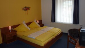 Une chambre d’hôtel avec un lit simple, une table de chevet, une chaise, un bureau avec un ordinateur, une fenêtre avec des rideaux et un élément de décoration murale.