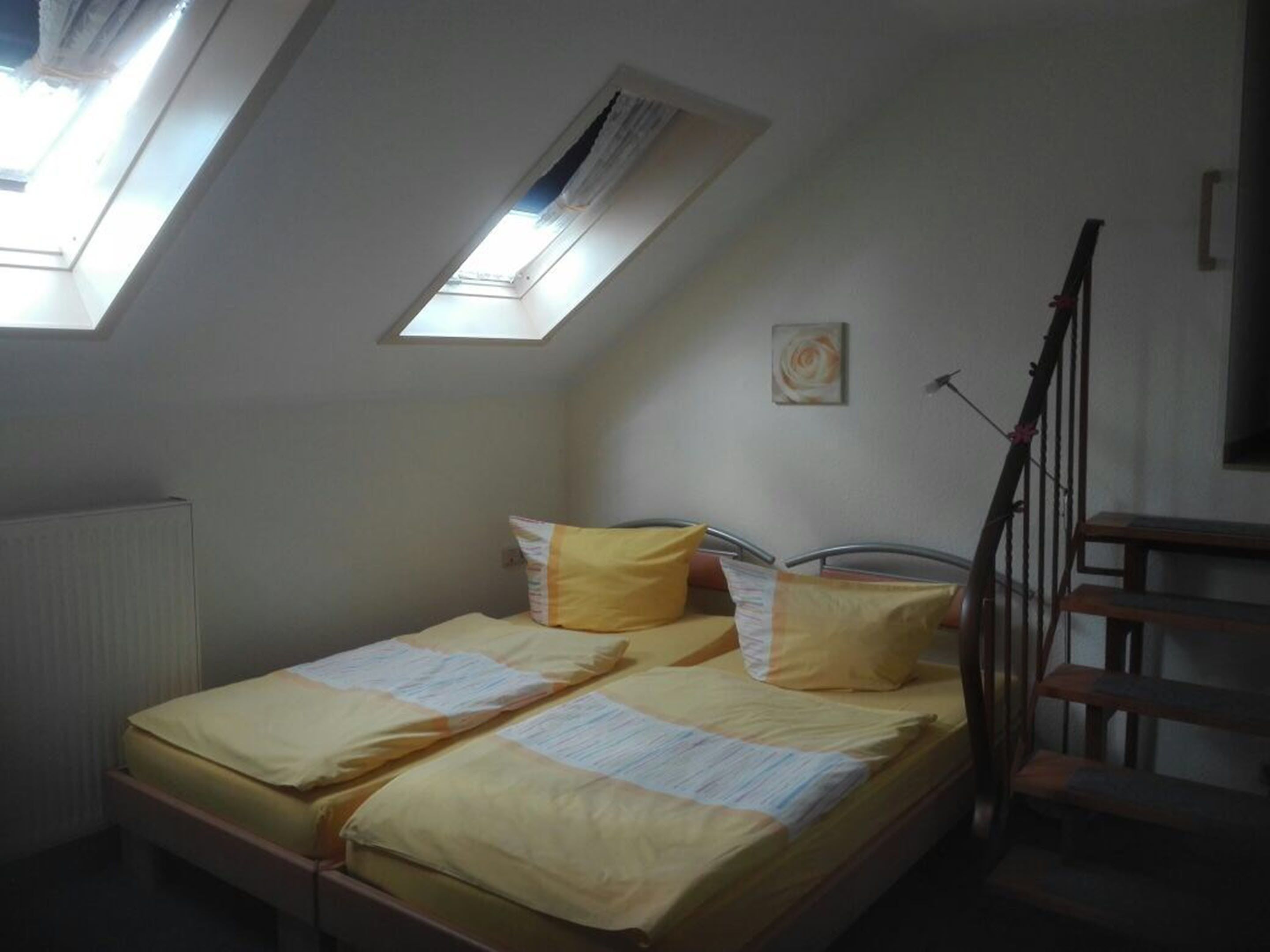 Ein kleines Zimmer mit einem Einzelbett, geneigter Decke und Oberlichtern, einem Heizkörper, einer Holztreppe und einem einzigen Wandbild.