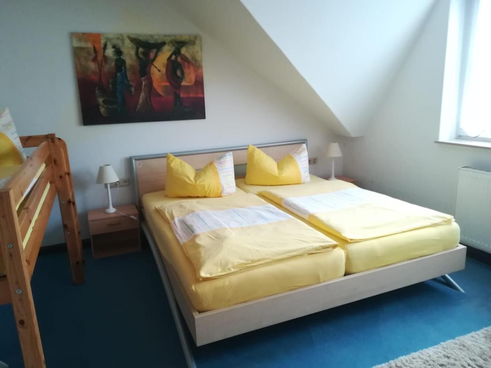 Una habitación con cama, mesita de noche, lámpara, un cuadro en la pared y ventana.