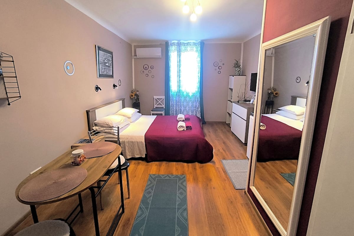 Een kleine, goed verlichte kamer met twee bedden, een bureau, een stoel, een spiegel en een raam met gordijnen met een patroon.
