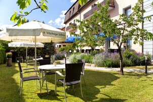 Un área de estar al aire libre con mesas y sillas, rodeada por un jardín cuidado y un edificio con balcones.