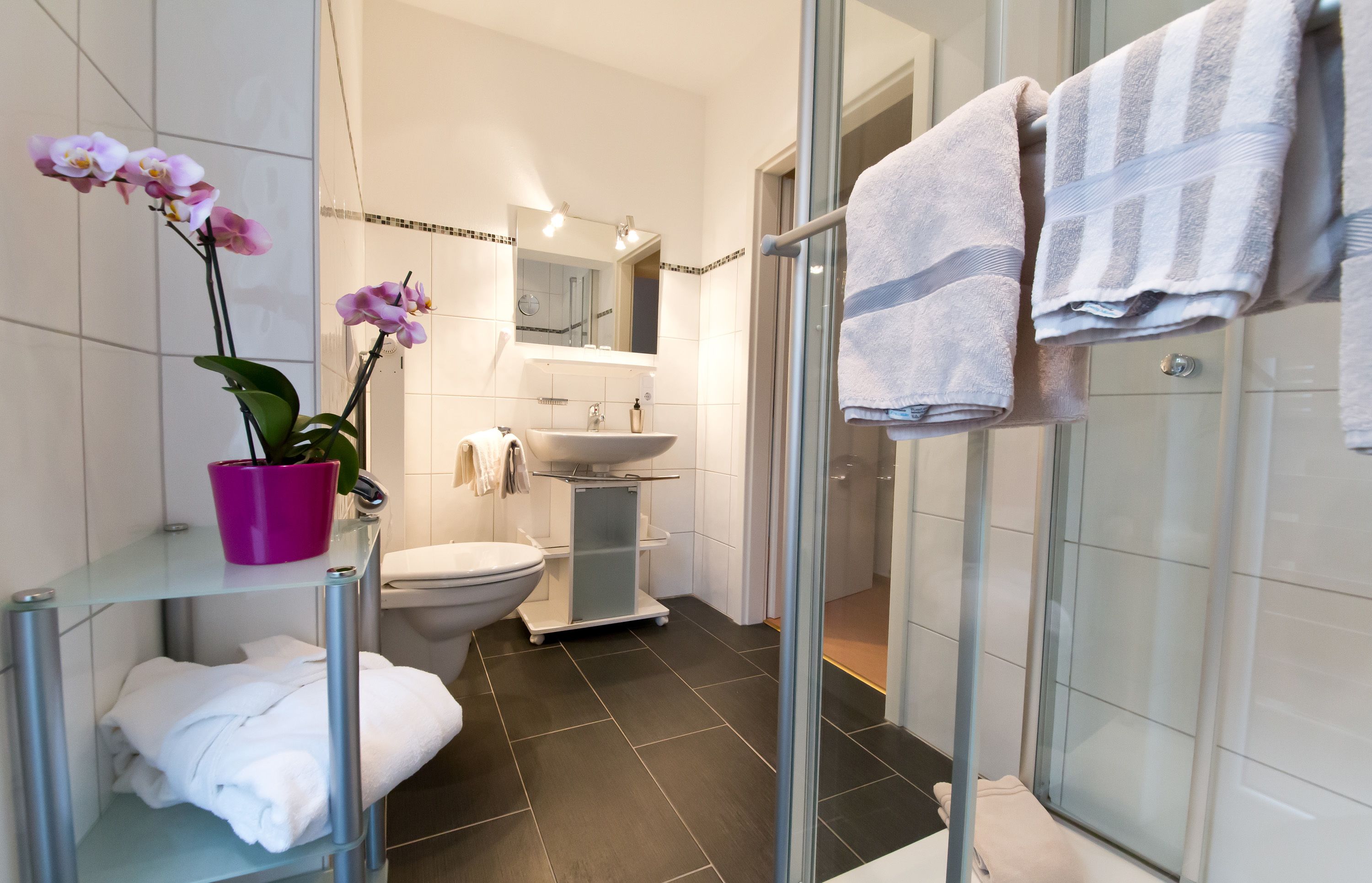 A modern bathroom with a glass shower enclosure, a white sink, and a toilet. There are white towels on a rack and a potted orchid on a shelf.