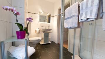 A modern bathroom with a glass shower enclosure, a white sink, and a toilet. There are white towels on a rack and a potted orchid on a shelf.