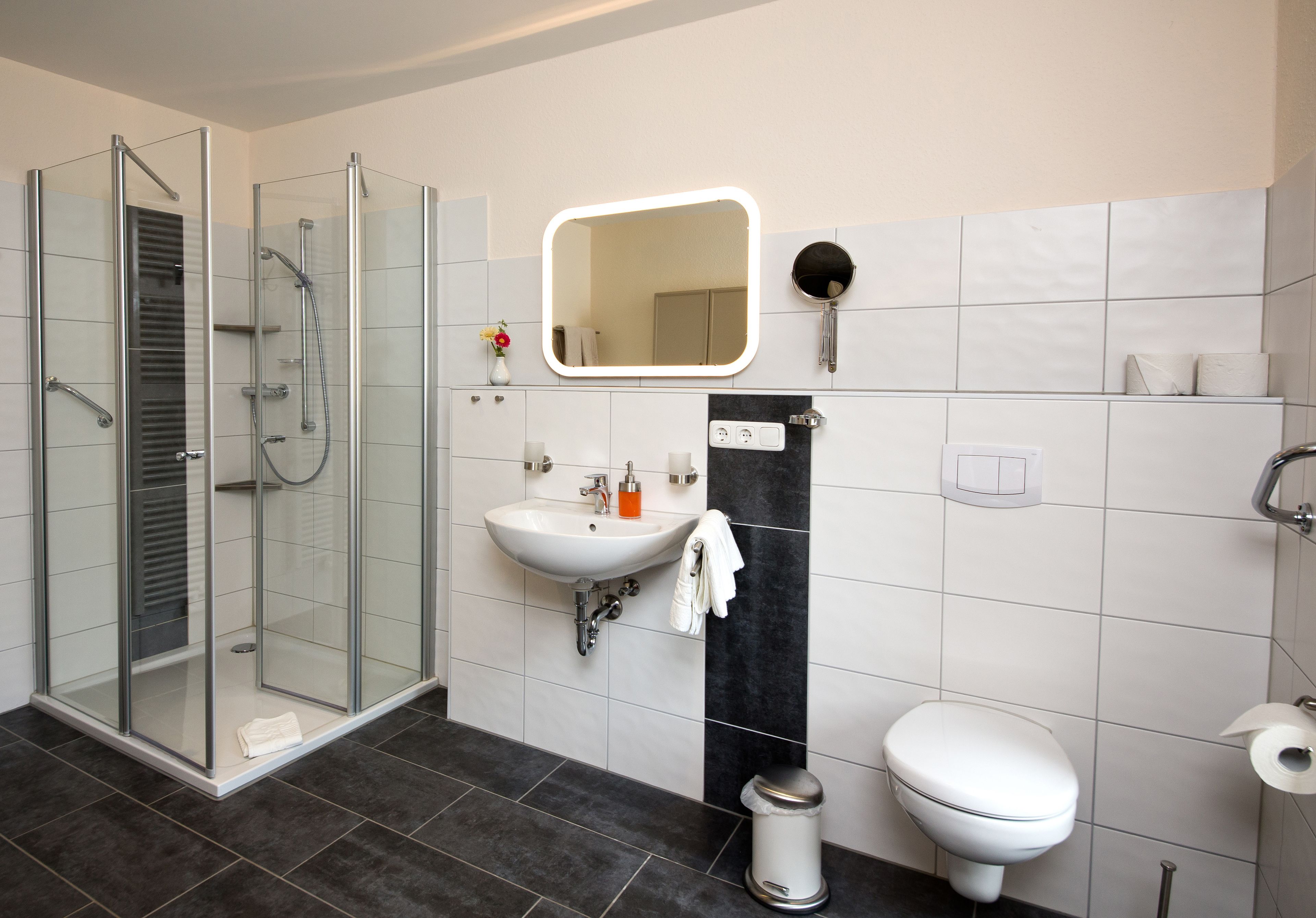Een moderne badkamer met een douchecabine van glas, een wit wastafelmeubel, een accentmuur in zwart, een toilet en een spiegel met ingebouwde verlichting.