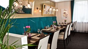 Een restaurant met smaragdgroene bekleden zitbanken, houten tafels met wijnglazen en hanglampen.