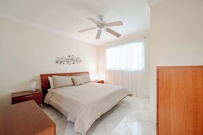 A bedroom with a bed, nightstands, a desk, a ceiling fan, a window with curtains, and a wall decoration.