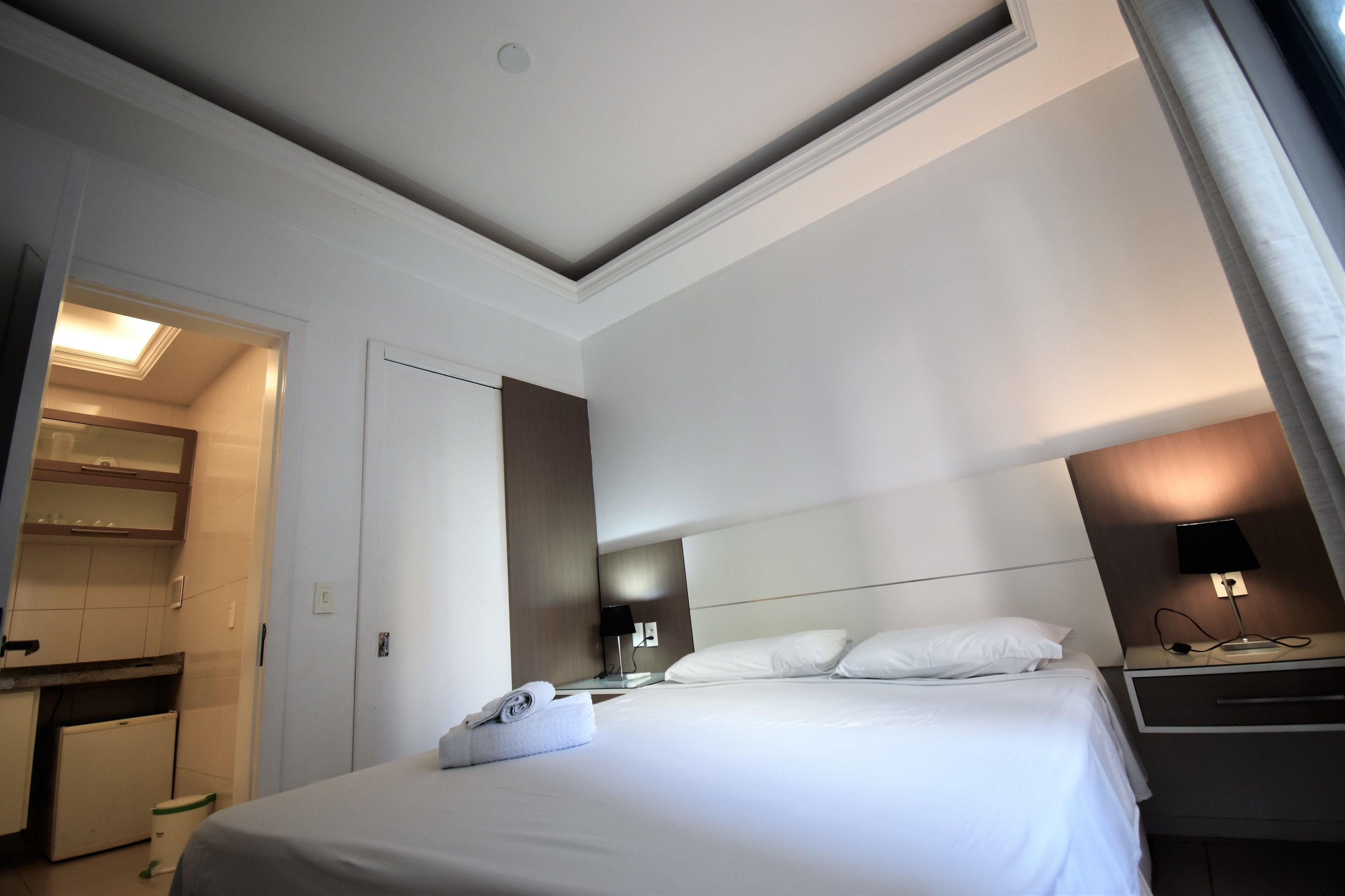 Habitación de hotel con una cama bien hecha, un escritorio pequeño con lámpara y vista al área de la cocina contigua.