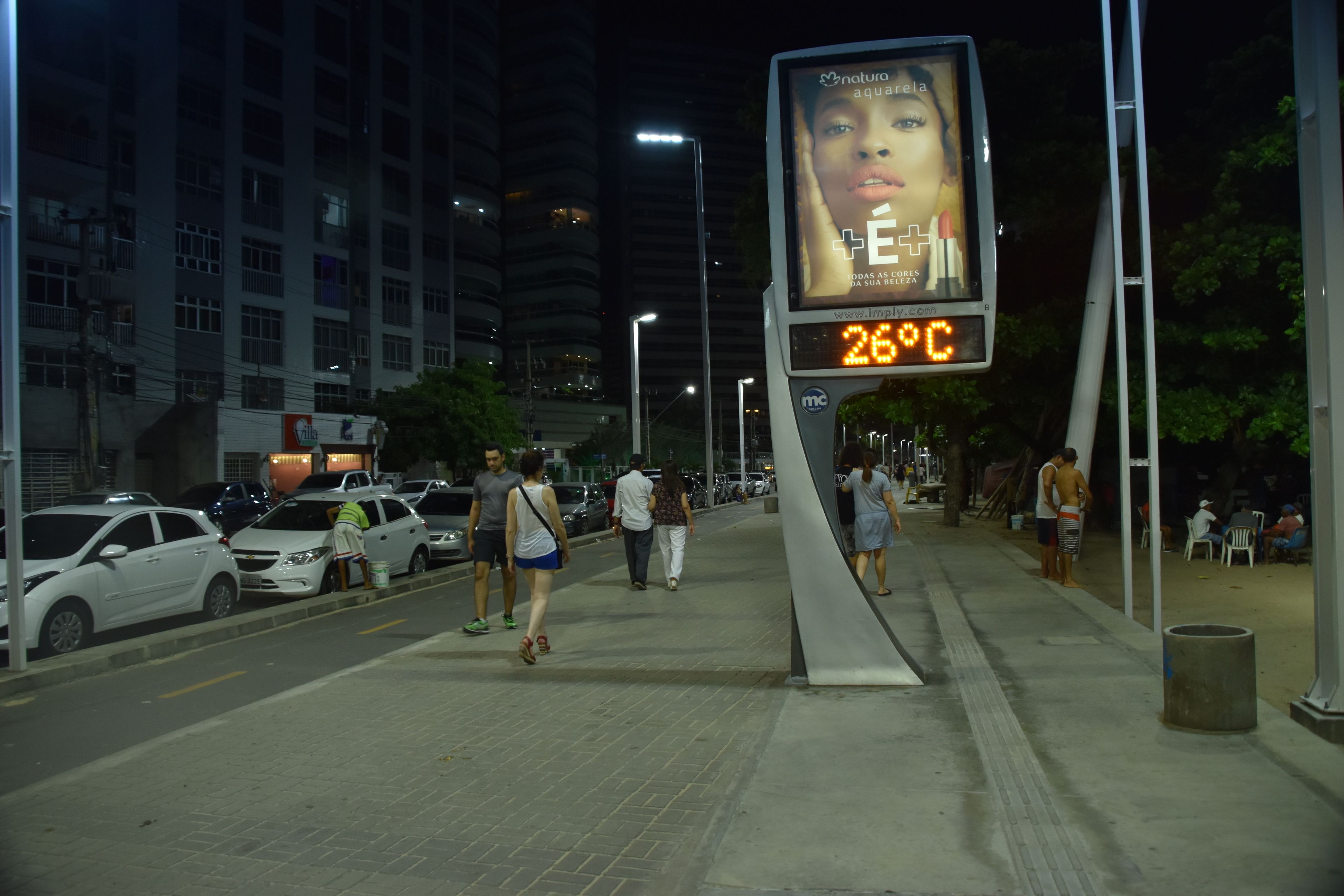 Cena noturna de rua com pedestres, carros estacionados e um visor digital de temperatura.