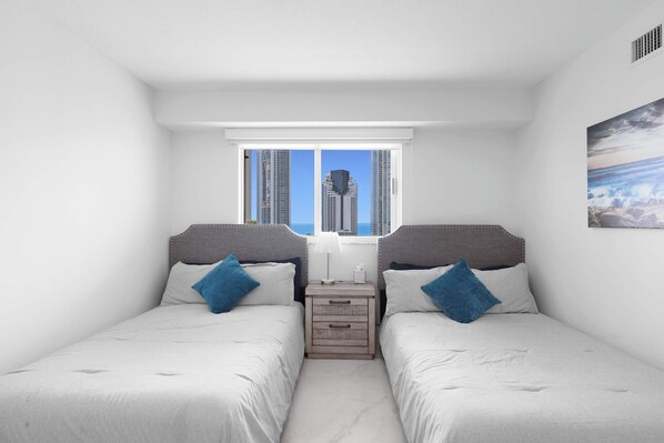 A bedroom with two beds, a nightstand, a window with a view of tall buildings, and a painting on the wall. - Le Marina Bay PH3 STR-03599 (Sunny Isles Beach)