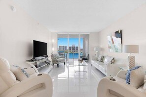 A modern living room with a flat-screen TV, a sofa, a coffee table, and a view of a cityscape through large windows. - Le Marina Bay PH4 STR-03600 (Sunny Isles Beach)