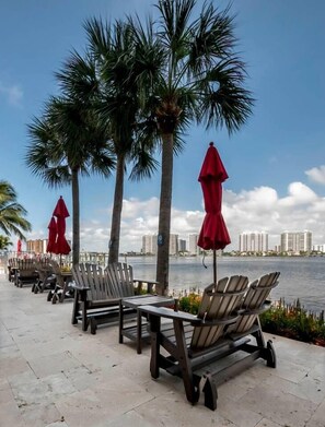 A waterfront patio with wooden chairs and red umbrellas, palm trees, and a view of high-rise buildings across the water. - Le Marina Bay PH4 STR-03600 (Sunny Isles Beach)