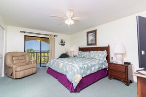 A bedroom with a bed, a recliner, a nightstand, a ceiling fan, and a view through a sliding door. - Panoramic Ocean Views at Ponce Landing 2 (St. Augustine)
