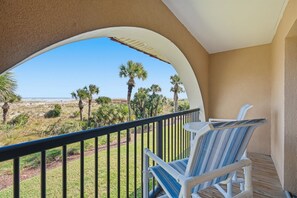 A balcony with two blue-striped chairs, a view of a beach with palm trees, and a clear sky.