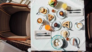 Ruokapöytä on katettu monenlaisilla aamiaisherkuilla, kuten leivonnaisilla, hedelmillä ja juomilla, ja sen ympärillä on punottuja tuoleja.