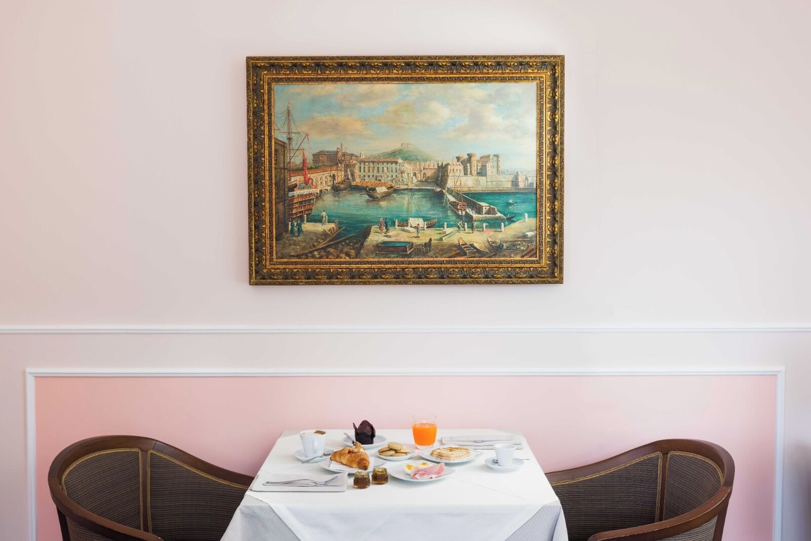 Zona pranzo con tavolo apparecchiato per la colazione, un quadro incorniciato raffigurante una veduta portuale e due sedie.