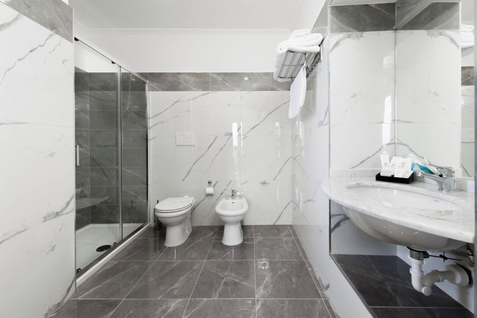 A modern bathroom with a glass shower enclosure, two toilets, a sink, and a towel rack.