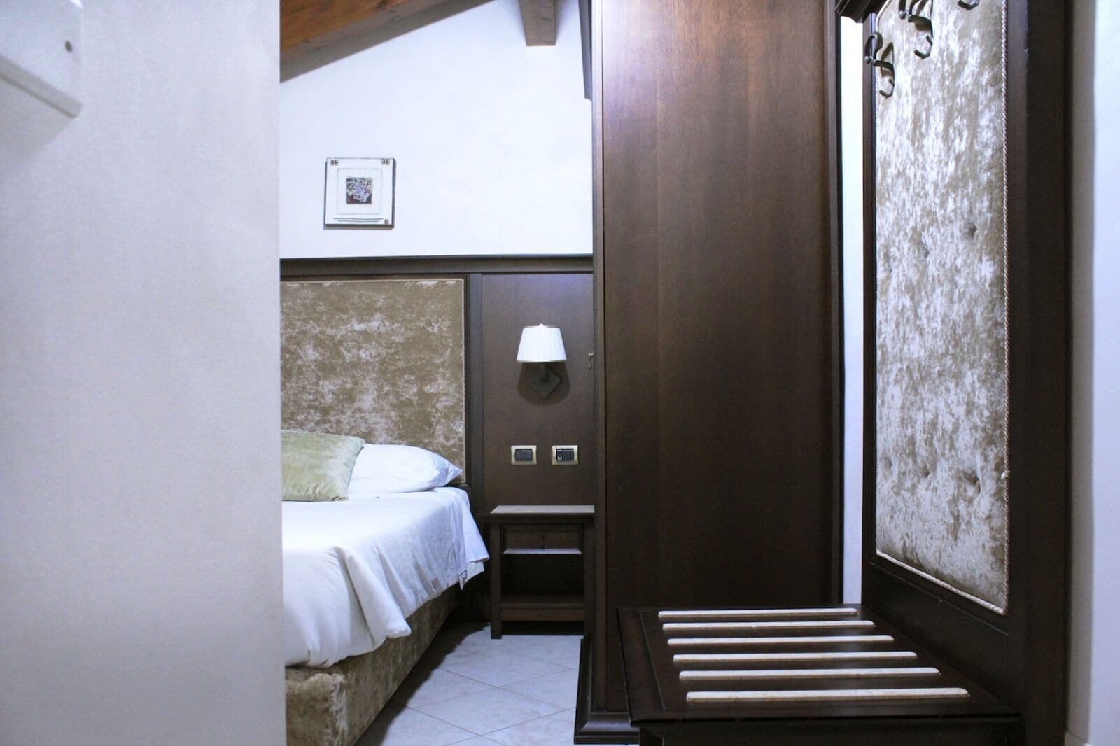 Une petite pièce étroite, comprenant un lit, une table de chevet, une lampe fixée au mur et un panneau mural texturé.
