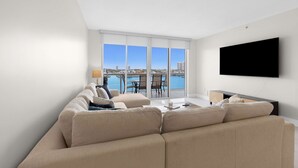 A modern living room with a large sofa, a flat-screen TV, and a view of a body of water through large windows. - Le Golden Bay 902 STR-00876 (Sunny Isles Beach)