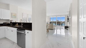 A modern kitchen with white cabinets, a black countertop, and a dishwasher. There is a dining area with a white table and chairs, and a view of a pool and cityscape through large windows. - Le Golden Bay 902 STR-00876 (Sunny Isles Beach)