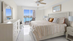 A bedroom with a bed, a dresser, a ceiling fan, a nightstand, a lamp, a chair, a window with a view of buildings, and a framed picture on the wall. - Le Golden Bay 902 STR-00876 (Sunny Isles Beach)