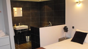 Una habitación de hotel moderna con una cama grande, una mesita de noche, una lámpara de pared y un baño con lavamanos y espejo.