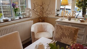 Una habitación acogedora con una silla blanca, una mesa con una taza y un platito, y una rama decorativa con luces.