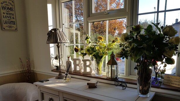 Una habitación con ventana, cómoda con cajones, lámpara y varios objetos decorativos, incluyendo flores y un cartel.