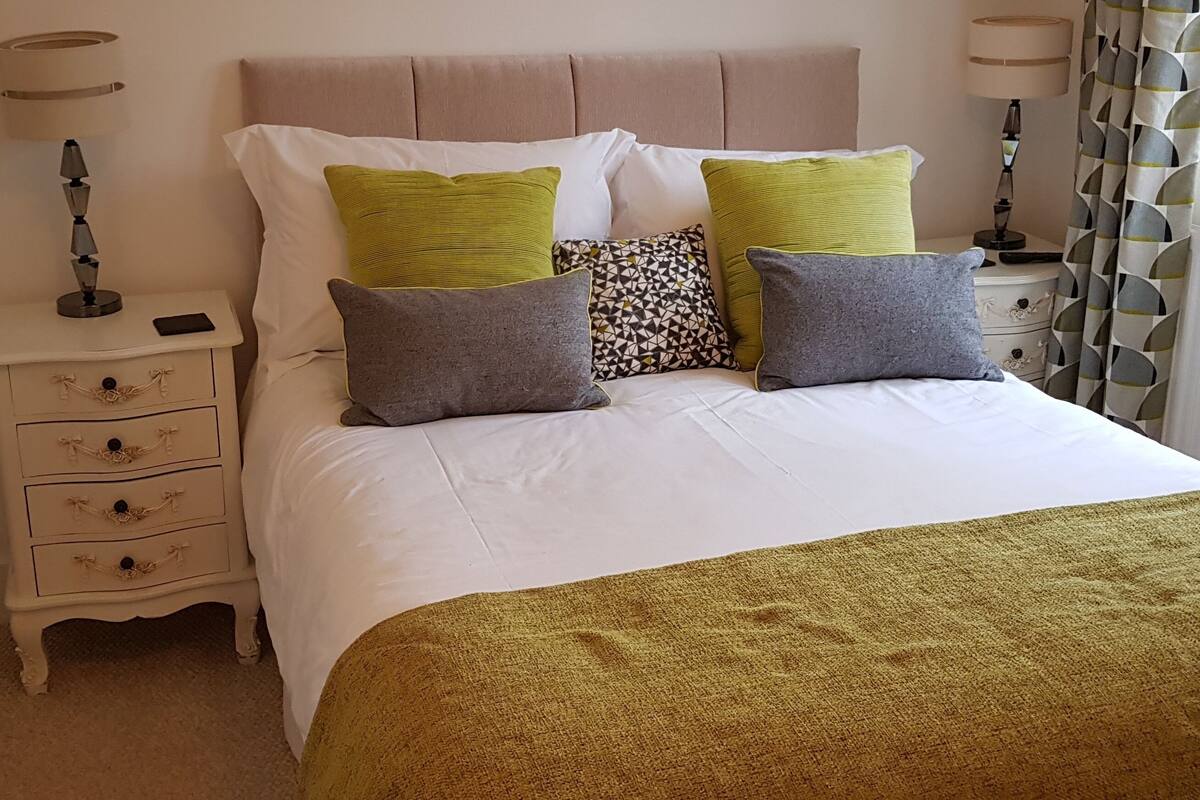 A neatly made bed with a white duvet and two green pillows, a patterned pillow, and a grey pillow. There is a bedside table with a lamp and a mobile phone on it. The room has a beige headboard and curtains with a geometric pattern.