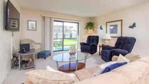 Un salon avec une table basse en verre, deux fauteuils, un canapé, un téléviseur et une vue sur une maison, visible à travers une porte coulissante.